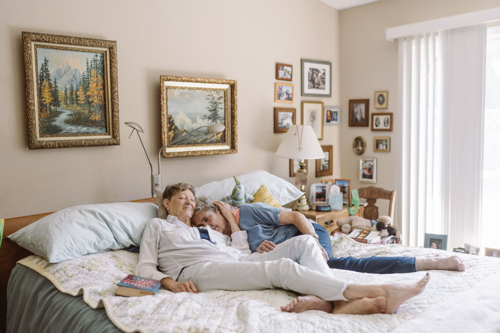 Two older women recline together on a bed in a cosy bedroom. They are embracing, eyes closed.