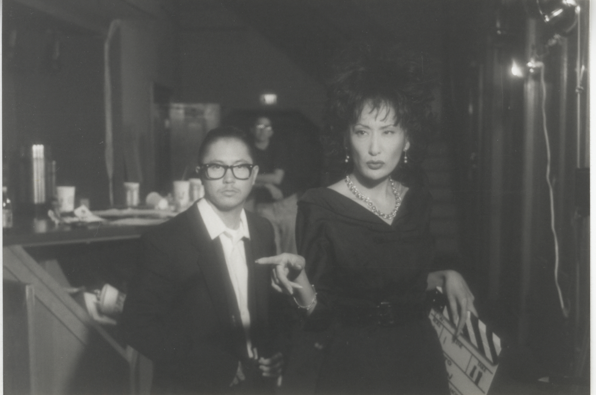 black-and-white image of person in glasses and suit, another person in a dress holding a clapper