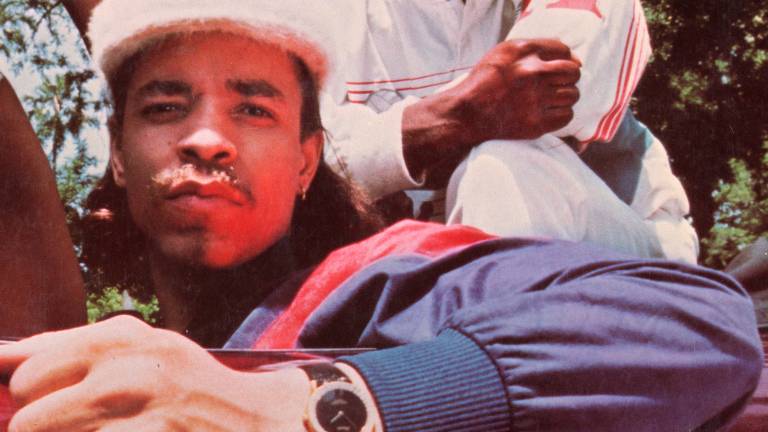 A man in a sports jacket and watch and white fuzzy kangol-style hat looks into the camera