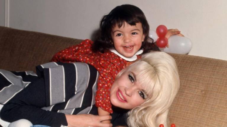 A beautiful white woman with blonde hair leans forward on a beige couch, a small white girl with brown hair, wearing a red dress, laughs while lying on the woman's back.