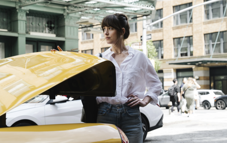A beautiful white woman with tied-back brown hair closes the trunk of a yellow cab in an urban street. She is wearing classic casual white shirt and blue jeans.