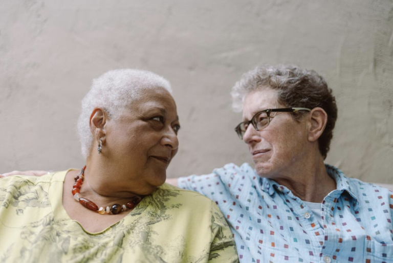 A Black woman with cropped white hair looks lovingly at a white woman with short grey hair and glasses, who is returning her gaze.