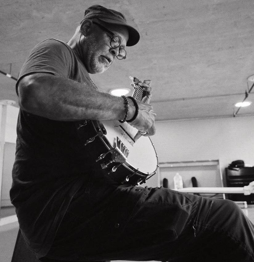 A candid black and white photo of a man with glasses and a hat playing a banjo. 