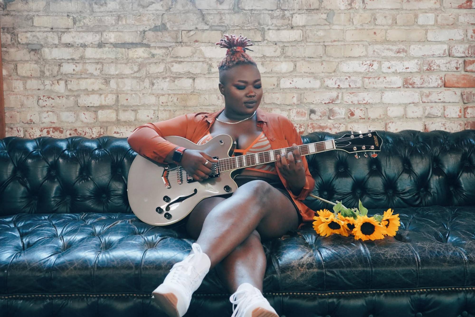 a young black woman in a yellow top sits on a couch with a guitar and some sunflowers against a white brick wall.