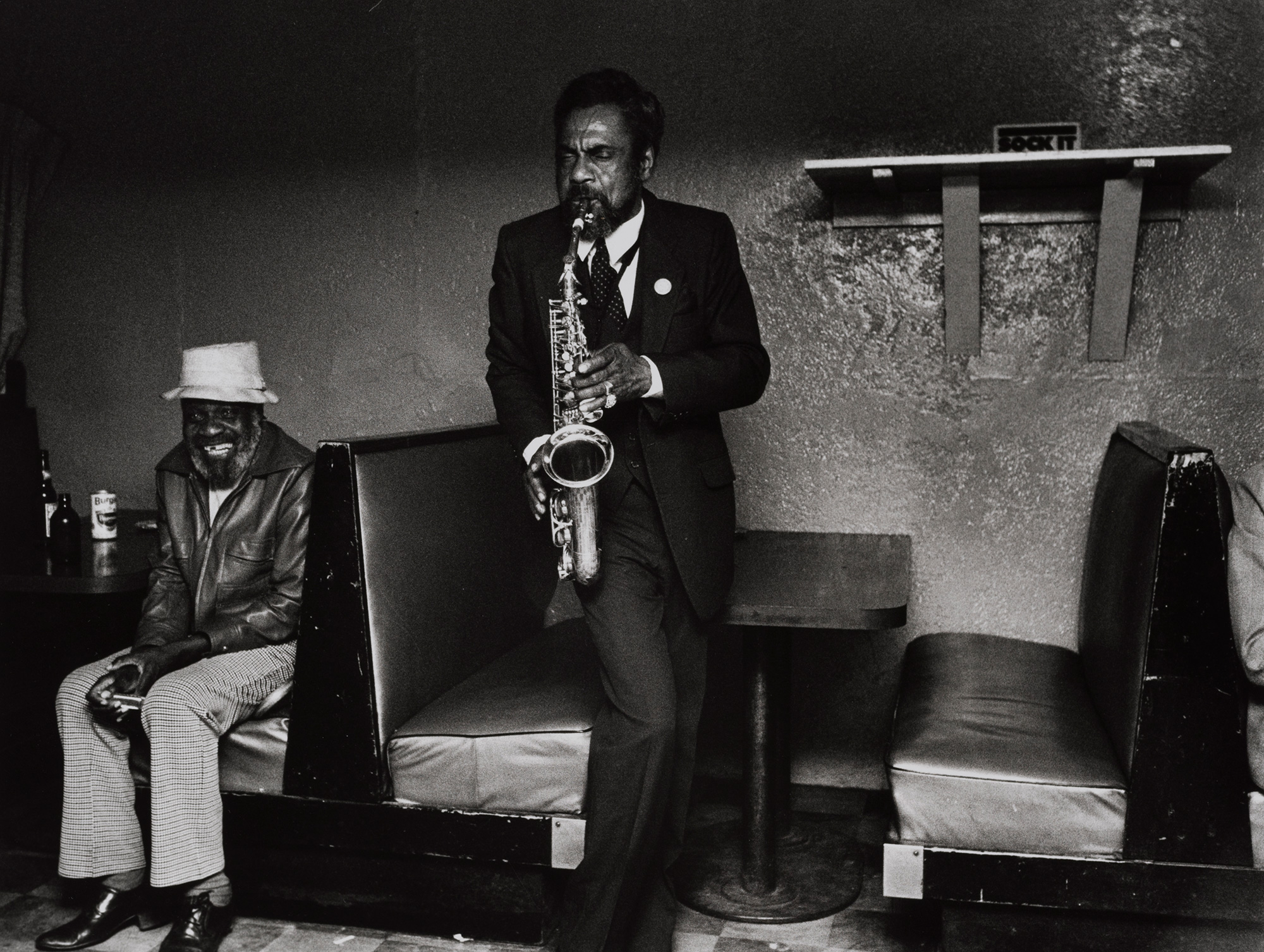A Black man with his eyes closed plays a saxophone in a dark nightclub while another man sits in a booth nearby, smiling
