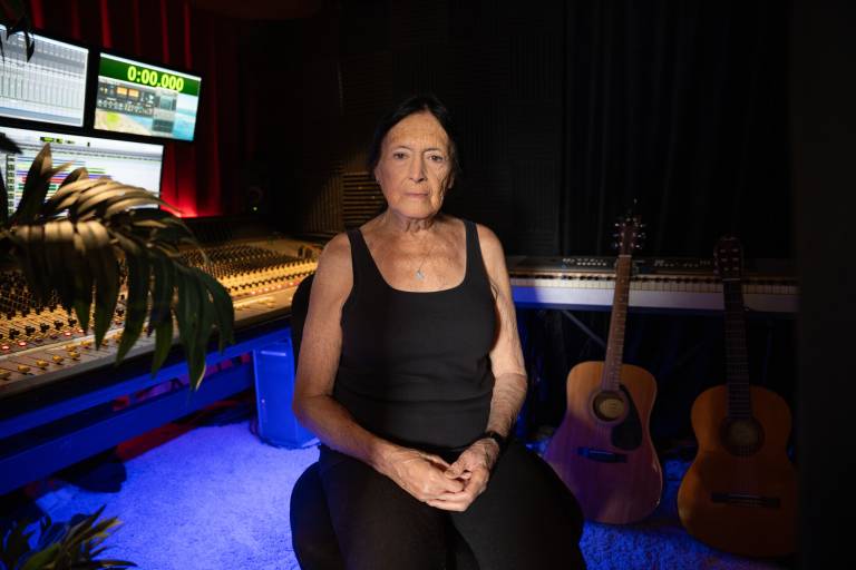 An older white woman dressed in black sits in a darkened room behind a music producer's desk alongside two acoustic guitars.