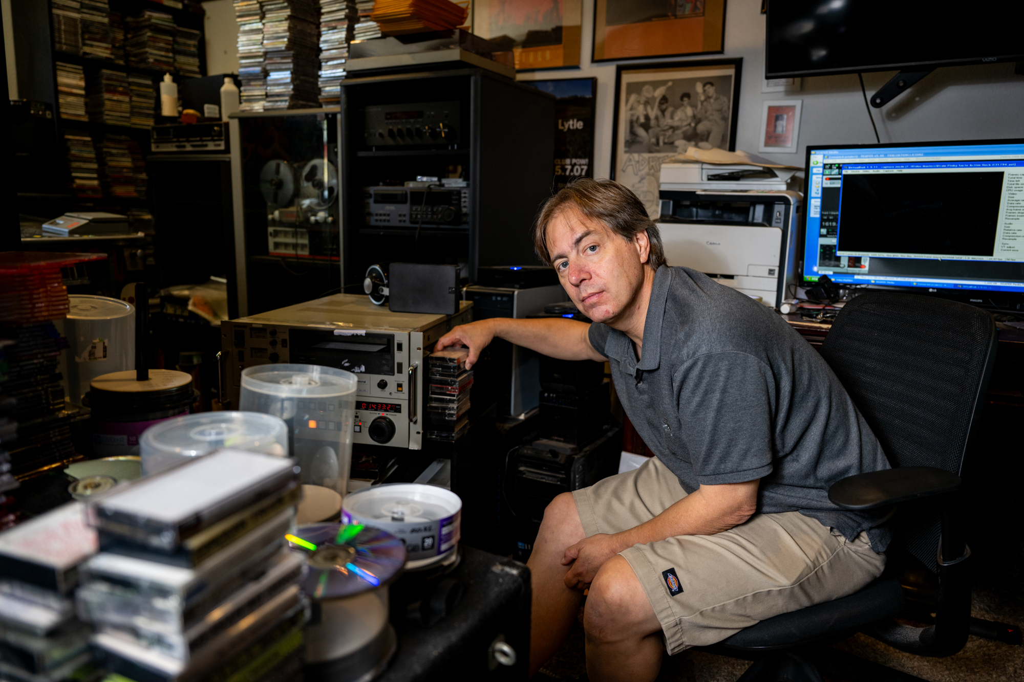 A man in his 50s wearing a polo shirt and beige shorts sits at a cluttered desk, his arm leaned upon a vintage analog tape machine