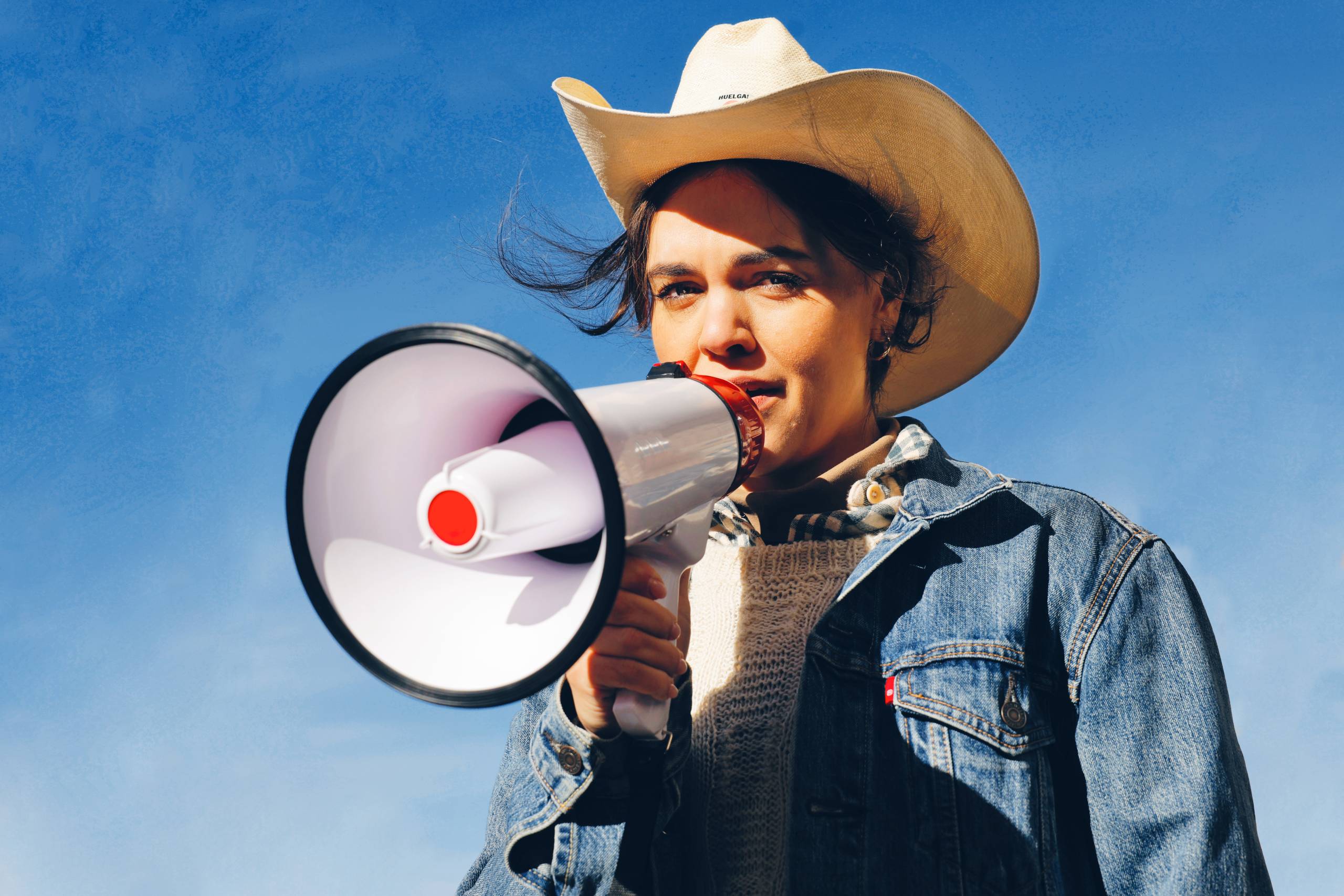 A Latina woman speaks into a bullhorn while wearing a cowboy hat.