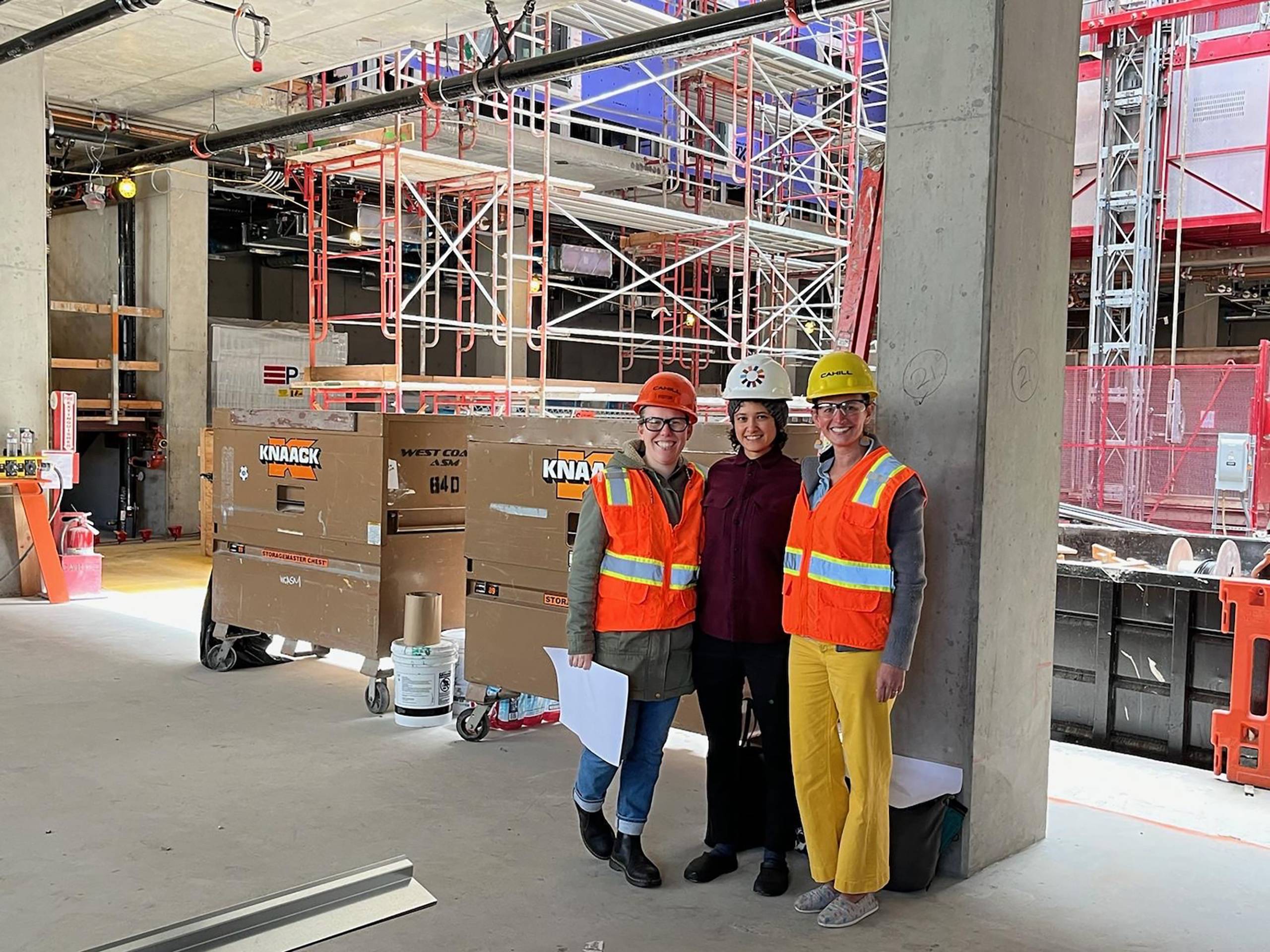 Three people in orange and yellow construction gear and helmets stand in front of a construction site with scaffolding and concrete