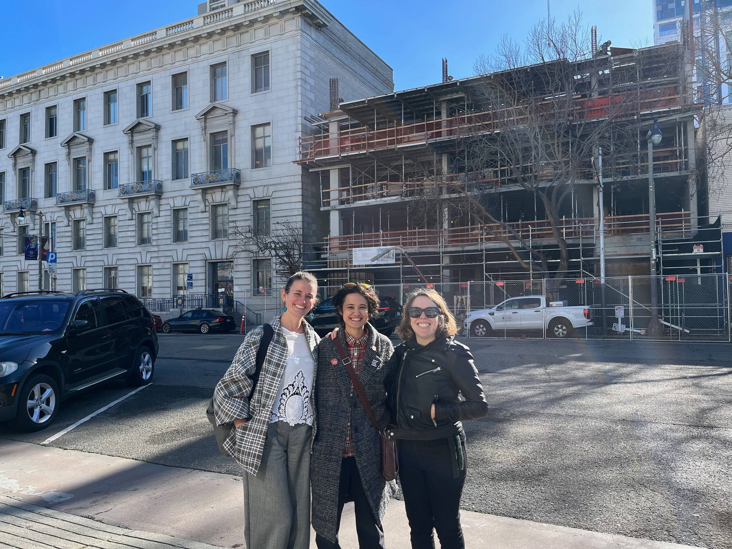 Three people stand on the sidewalk across the street from a building, under construction, in the background, posing for the camera