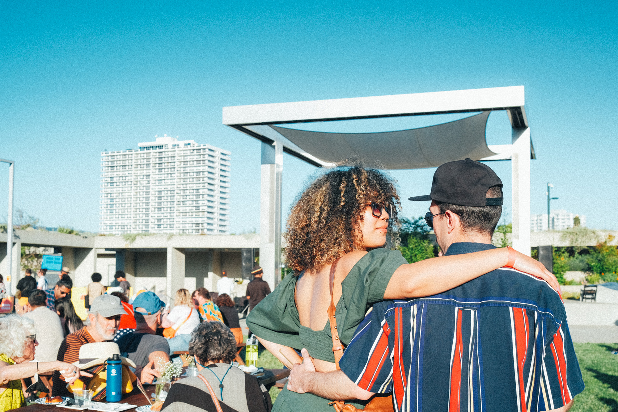 A crowd picnicking on a sunny day, including a smiling couple with arms around each other's shoulders