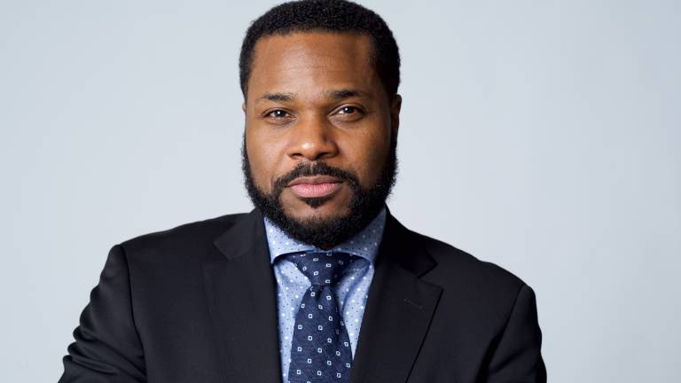 A black man in a beard and mustache wearing a stylish suit looks at the camera against a very light blue backdrop
