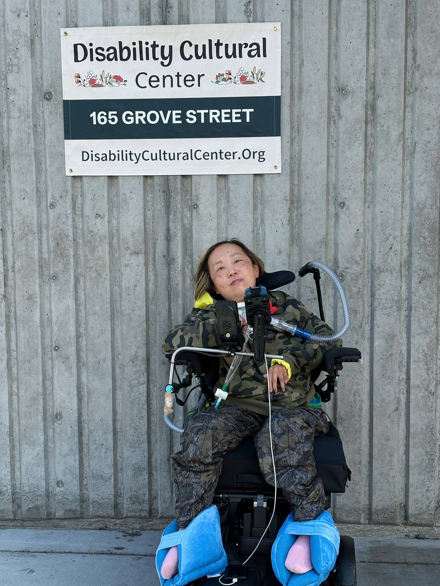A person in a wheelchair looks at the camera in front of a concrete wall with a sign reading "Disability Cultural Center, 165 Grove Street, disabilityculturalcenter.org."