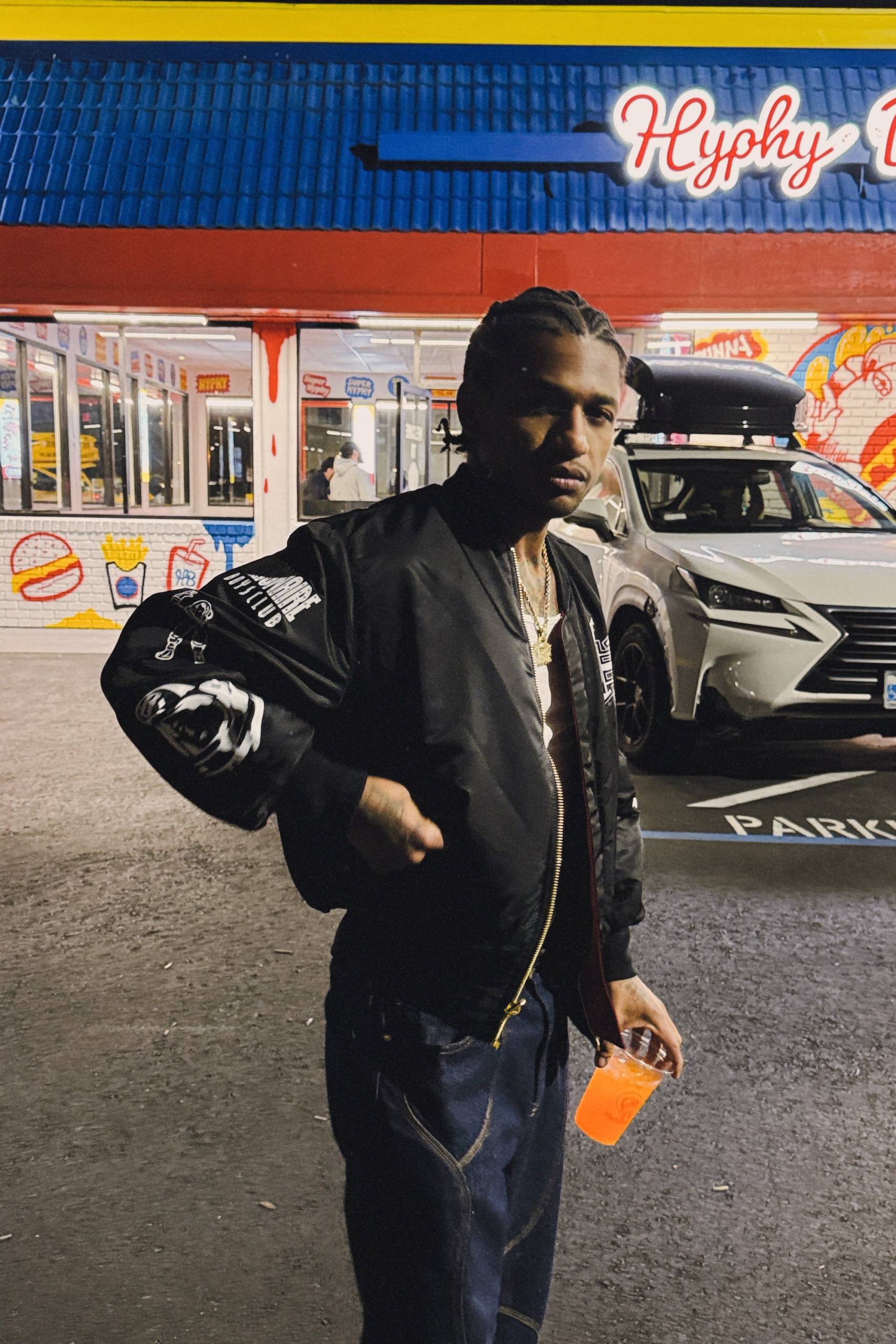 A man in a black jacket and braids poses for a photo outside of a burger joint while holding a soda. 