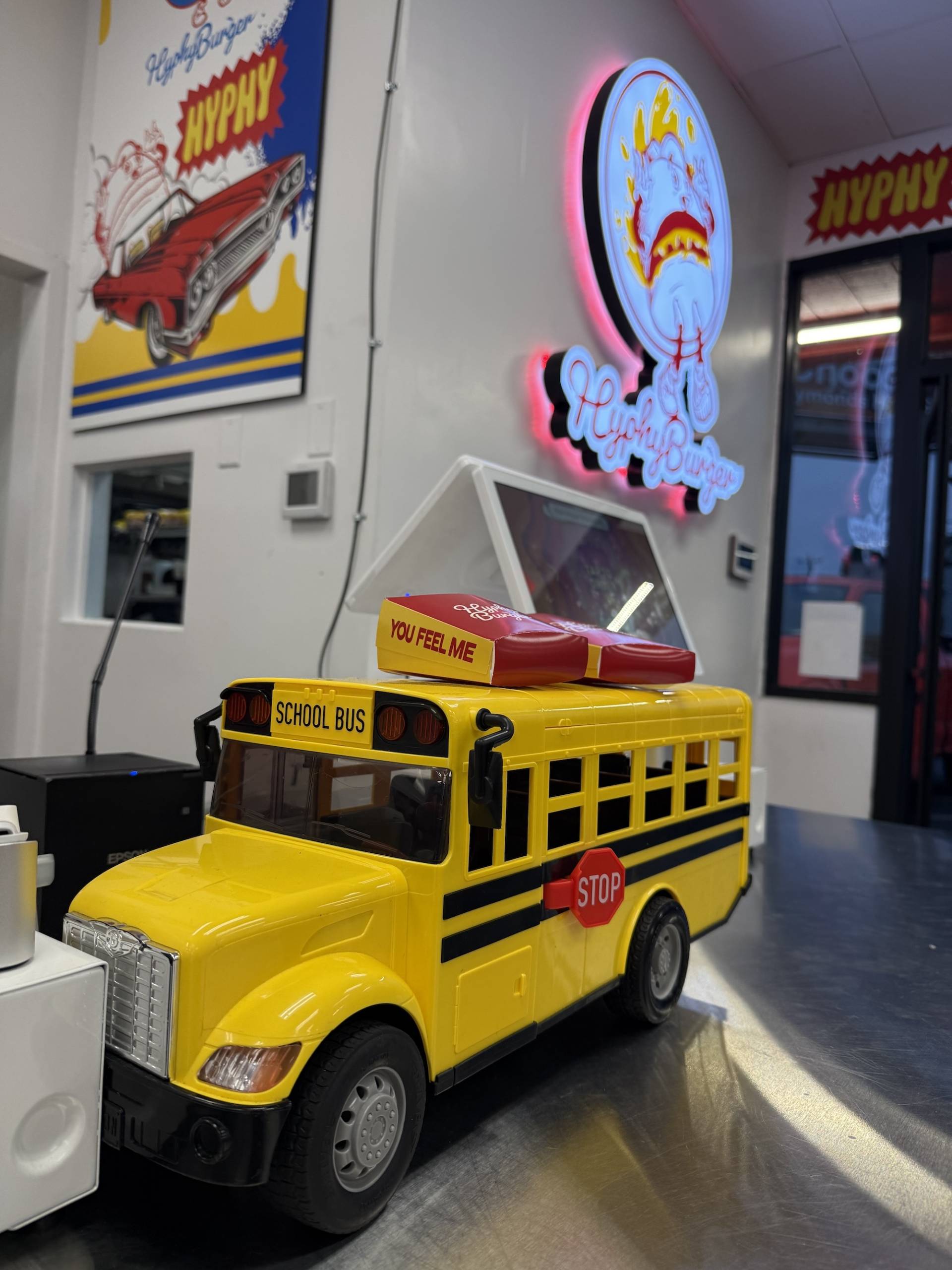 A mini yellow bus and signage that reads "you feel me" appear at the Hyphy Burger kiosk.