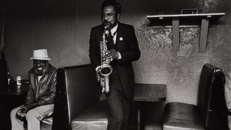 A Black man with his eyes closed plays a saxophone in a dark nightclub while another man sits in a booth nearby, smiling