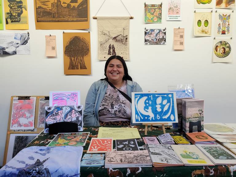 A woman sites at a table full of small printed matter, in front of a wall covered in hanging printed matter