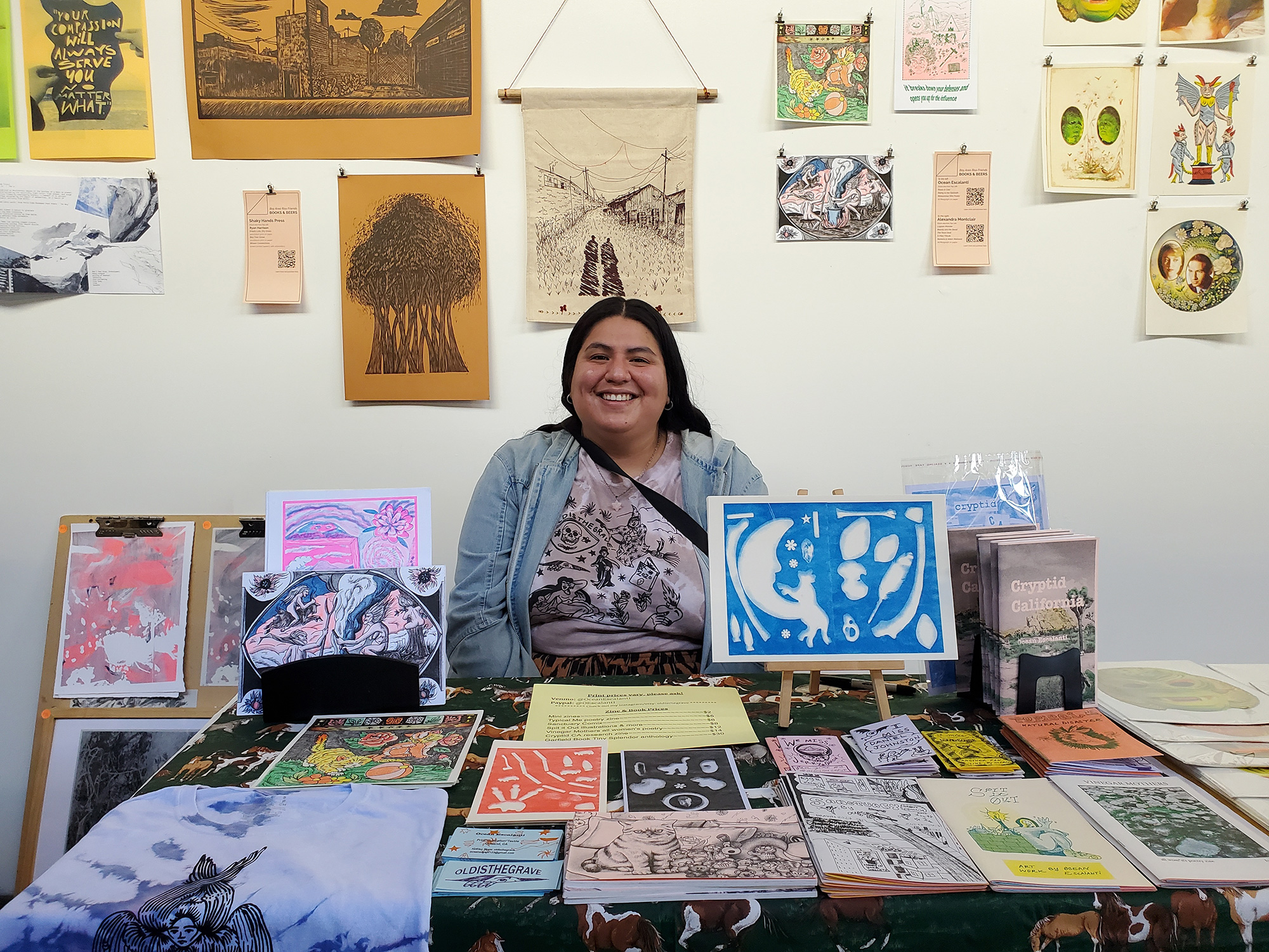 A woman sites at a table full of small printed matter, in front of a wall covered in hanging printed matter
