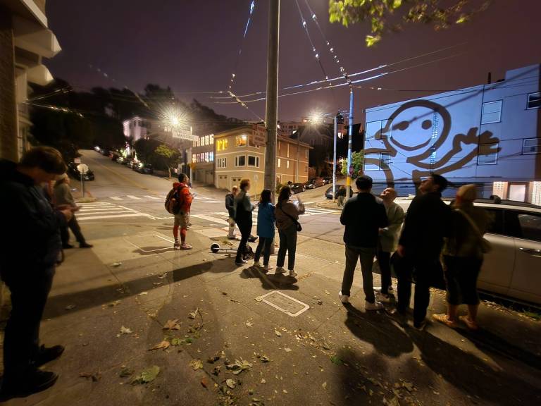 A group of people gather on a street corner at night, opposite a residential building. A projection of a simple black and white figure waving is being projected onto the side of the building.