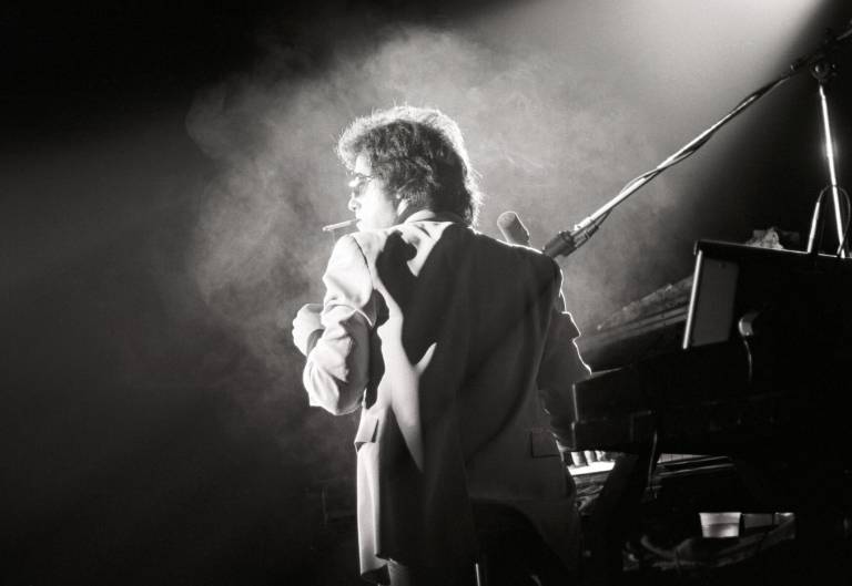 Billy Joel performing at his piano, on stage, cigarette in mouth, in front of a dry-ice filled spotlight. The photograph is black and white.