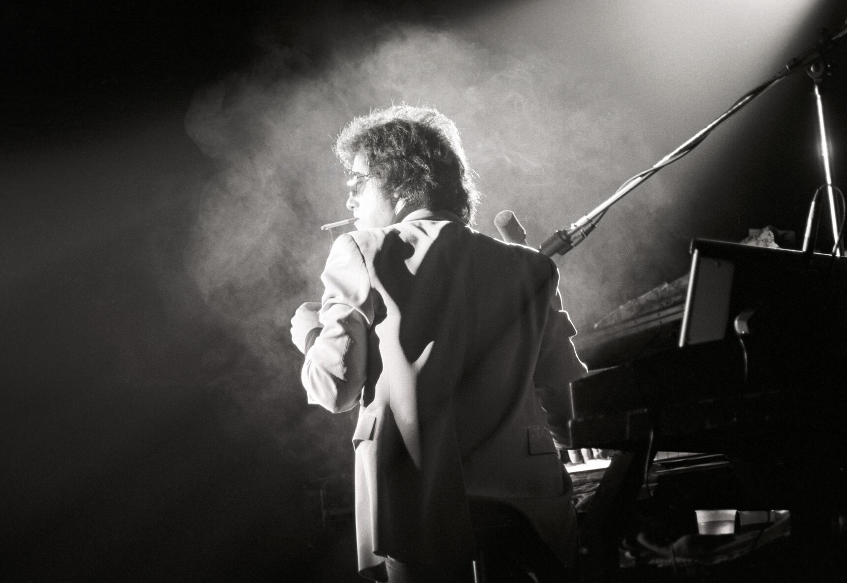 Billy Joel performing at his piano, on stage, cigarette in mouth, in front of a dry-ice filled spotlight. The photograph is black and white.