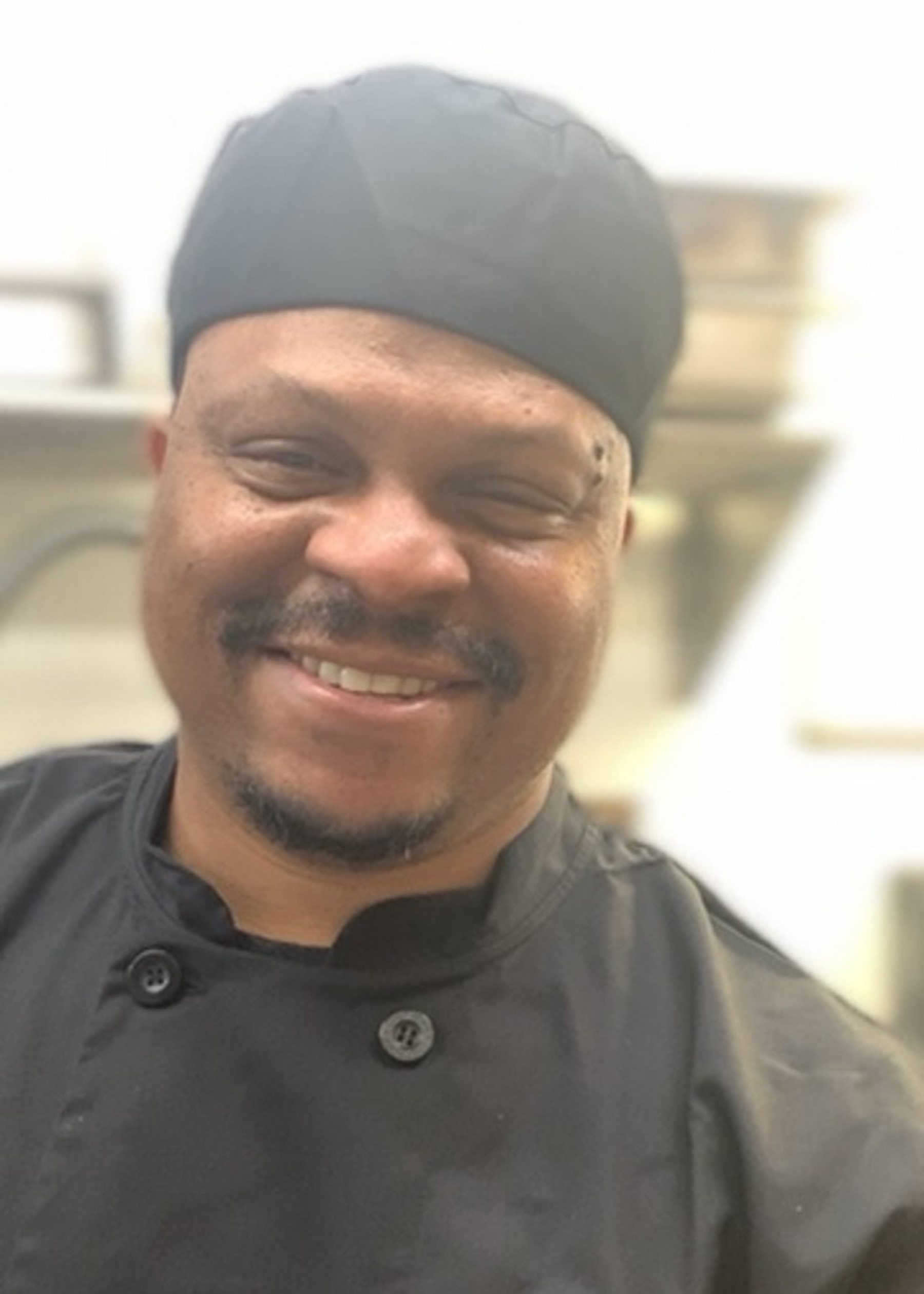 A middle-aged Black man in a black chef's coat and chef's hat smiles for a portrait.