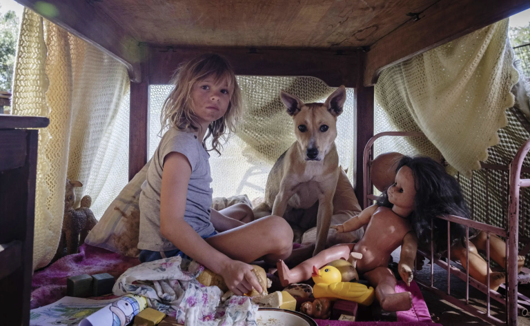 A little blonde white girl with a dirty face sits under a table with her dog, doll and some rags.