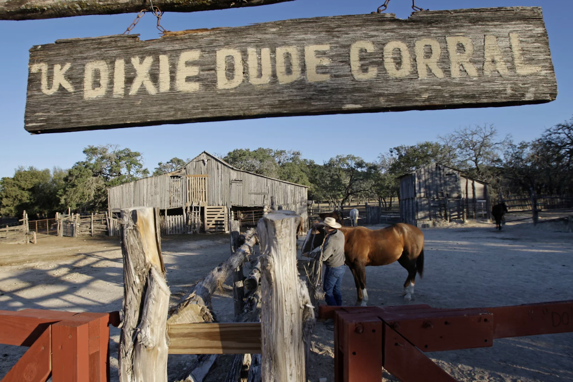 An aged wooden sign reading 'Dixie dude corral' hangs over a yard and stables where a man in a cowboy hat tends to a brown horse.