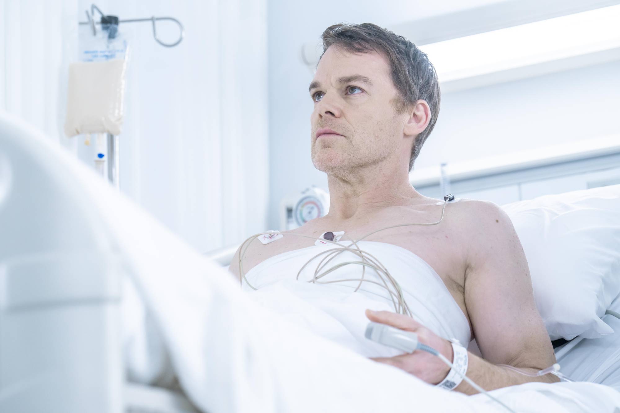 A shirtless white man leans upwards to an almost-seated position in a hospital bed. The room is brightly lit.