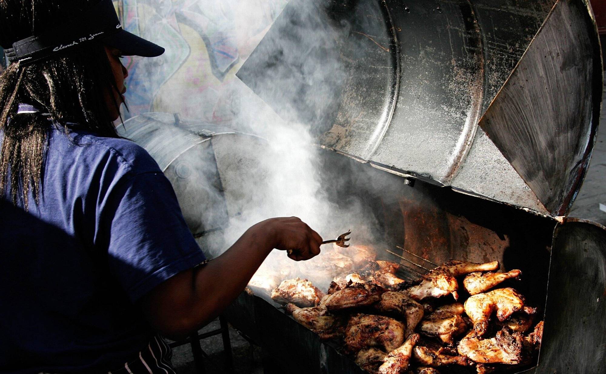 A Black woman wearing long braids, a purple t-shirt and a baseball cap holds a three-pronged fork over an open grill of smoking chicken pieces.