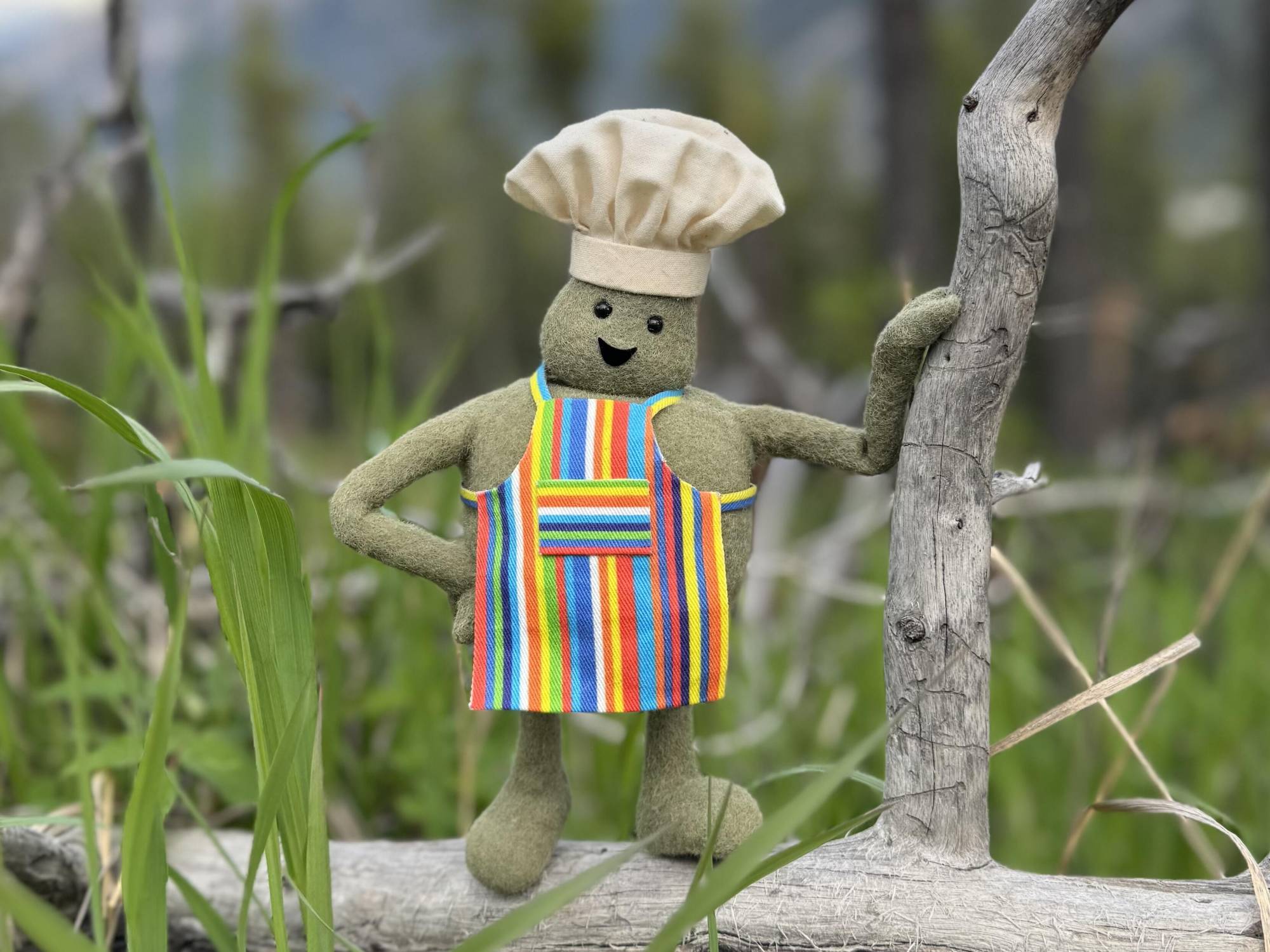 A tiny green puppet wearing a striped apron and chef's hat, smiles, posing next to a plant and blades of grass.