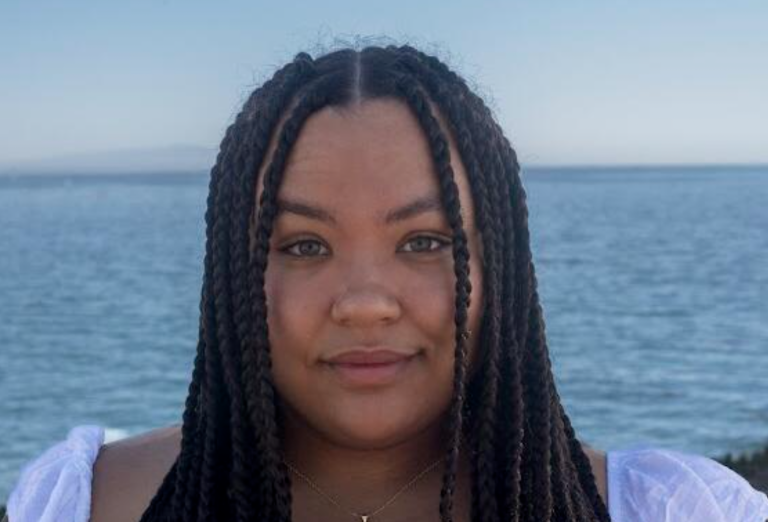 A young Black woman with long flowing braids stands facing the camera, the ocean stretched out behind her.