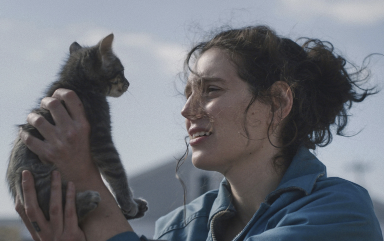 A 20-something white woman with dark messy hair tied back, stands in the street holding up and intently staring into the face of a kitten.