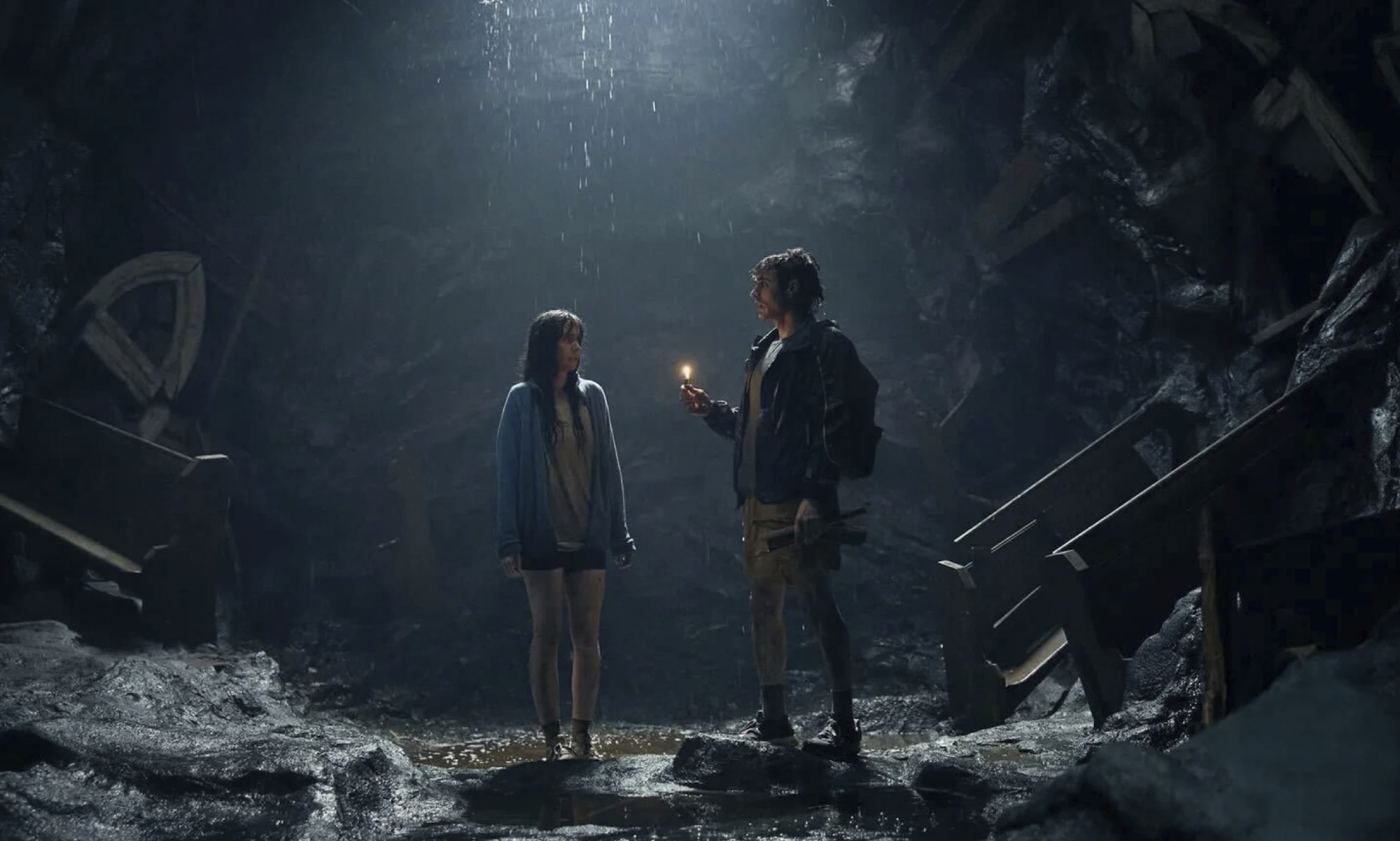 A woman and man, both wet and disheveled stand in an underground cave, surrounded by detritus.