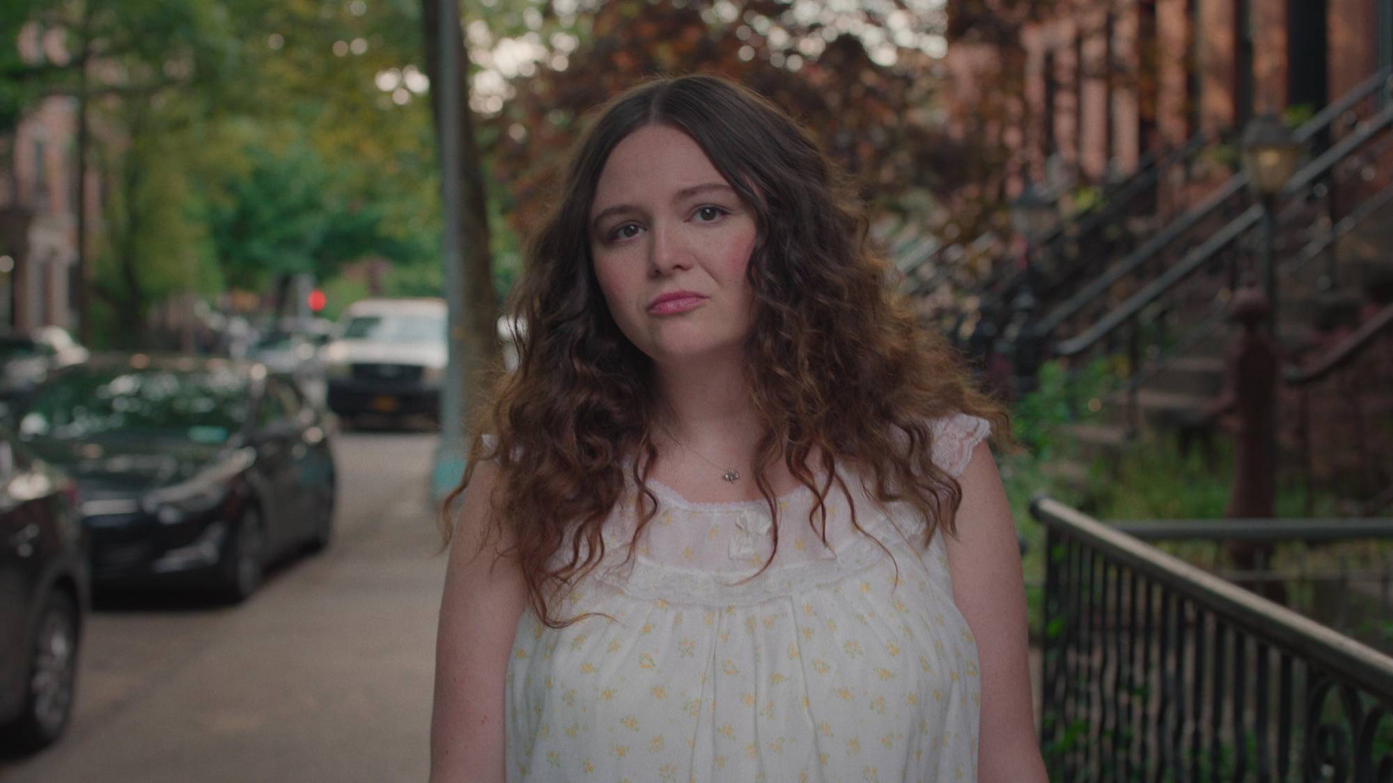 A fresh-faced white woman with shoulder length brown hair walks through a New York street, trees and brownstones visible behind her.