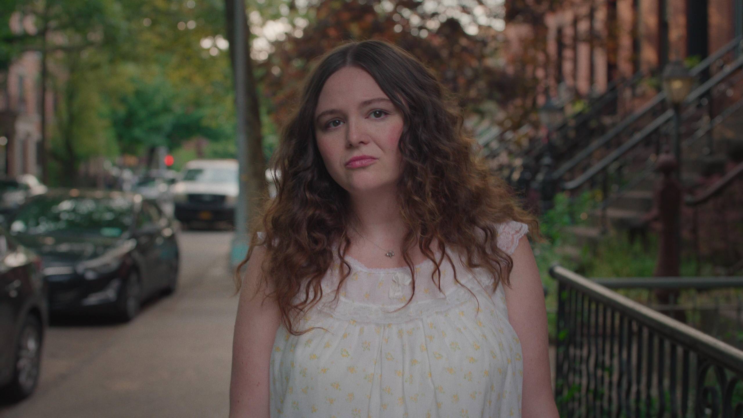 A fresh-faced white woman with shoulder length brown hair walks through a New York street, trees and brownstones visible behind her.