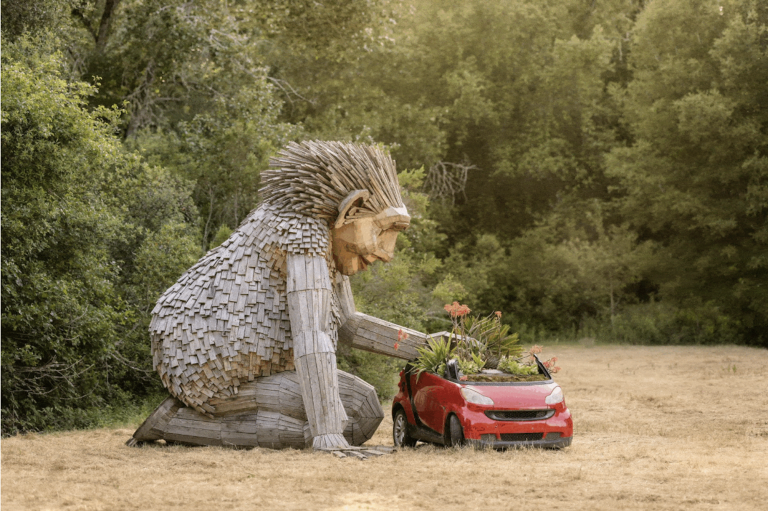 A giant sculpture of a troll, made out of wood fragments, kneels on the ground in lush, outdoor surroundings, appearing to place plants inside a red car.