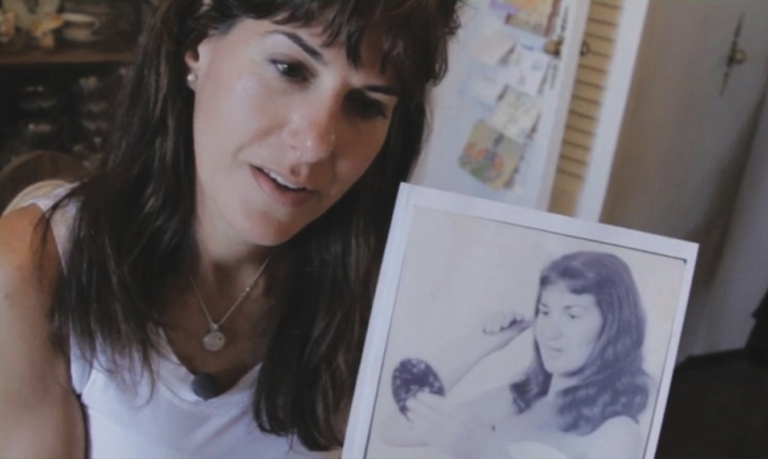 A white women with long brown hair, wearing a white t-shirt, holds up a black and white photograph of a similar looking woman from a different era.