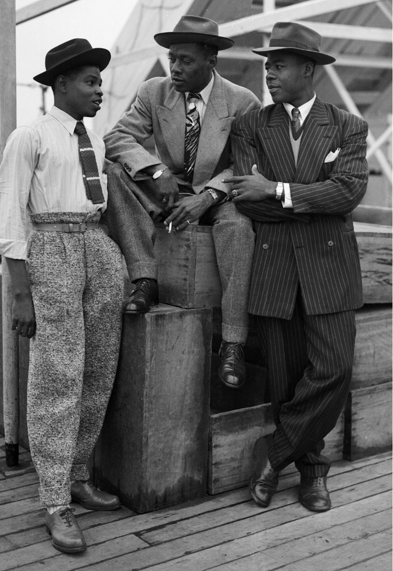 Three dapper Black men pose on a dock wearing smart, 1940s-era suits, ties and hats.