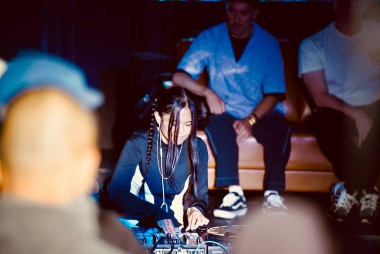 A woman, head down, looking at turntables as she DJs.