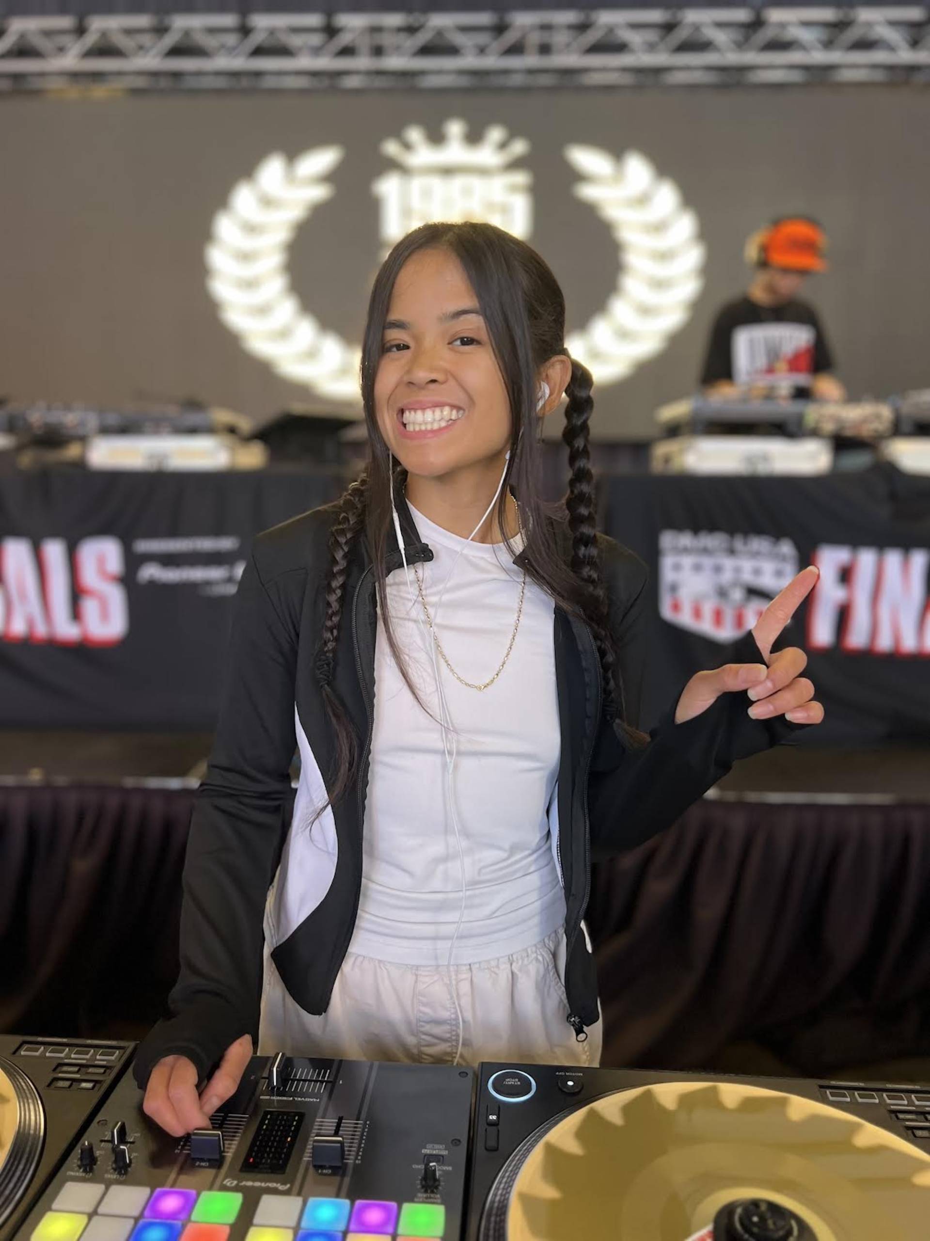 A woman with long black hair, a white shirt and a black jacket stands behind a DJ table and looks directly into the camera while smiling.