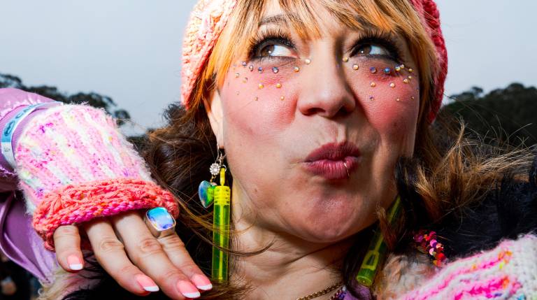A woman puckers her lips while showing off pink and red knit gloves and hat, wearing earrings and small sparkly adornments on her face
