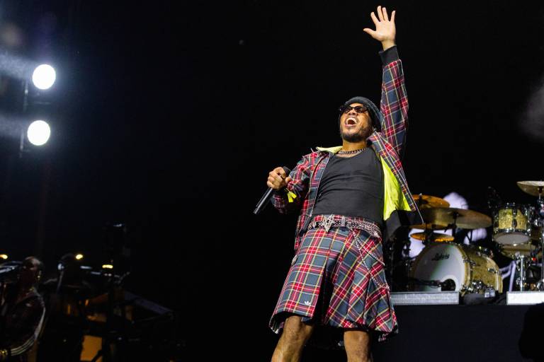 A Black man in sunglasses, a black top and a plaid kilt with matching jacket raises his left arm in the air while holding a microphone with his right