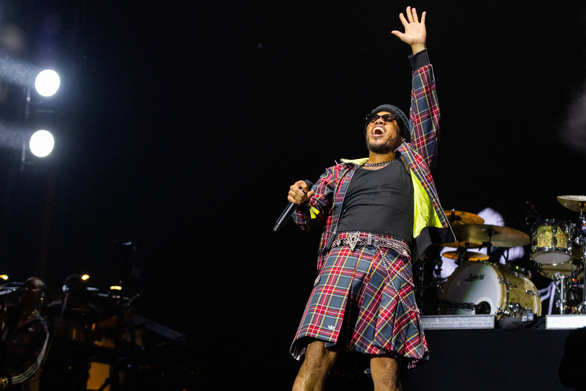 A Black man in sunglasses, a black top and a plaid kilt with matching jacket raises his left arm in the air while holding a microphone with his right