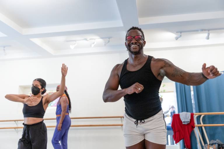 A Black man in black tank top and white shorts dances in a rehearsal space with white walls and white ceiling, as other dancers move in the background.