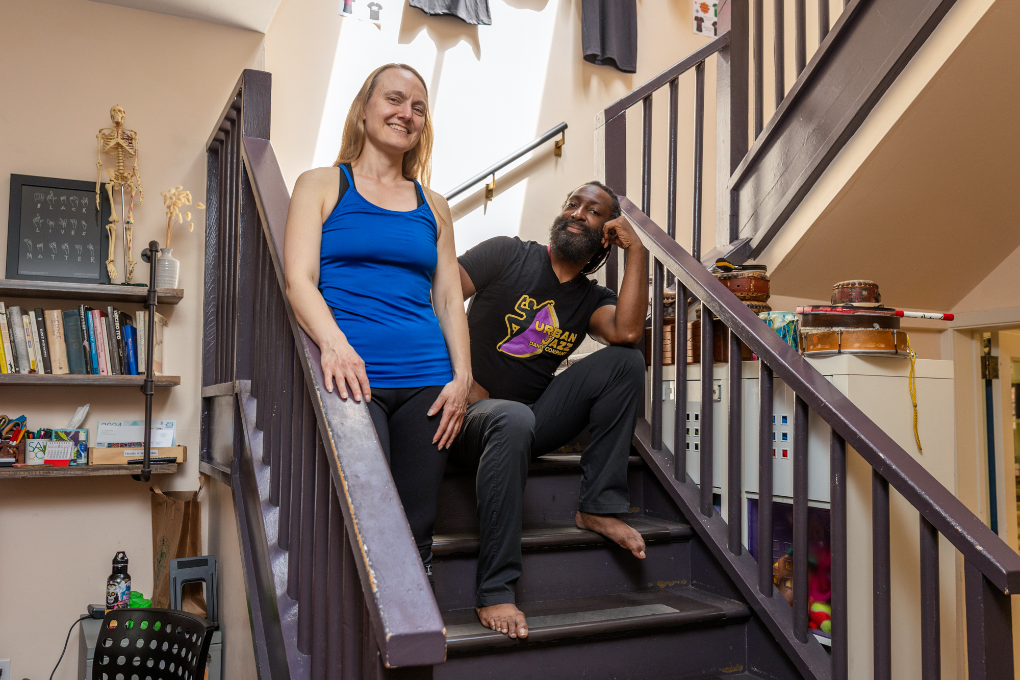 A white woman in a blue tank top and a Black man in jeans pose on a dark wooden stairwell, looking at the camera