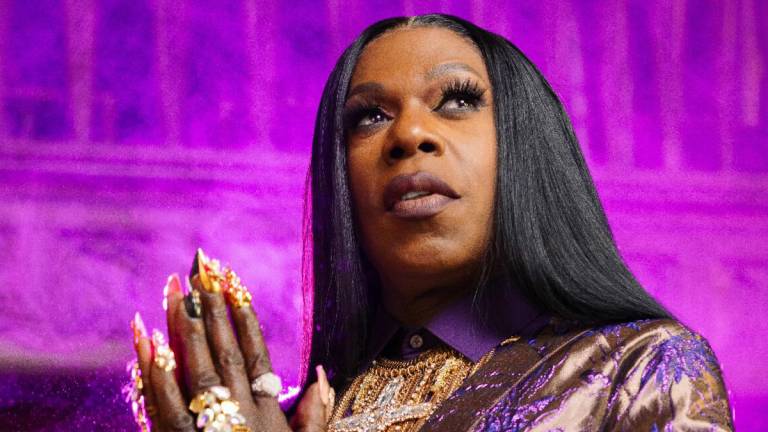 Big Freedia holds her hands together in prayer in a church illuminated by a purple glow.