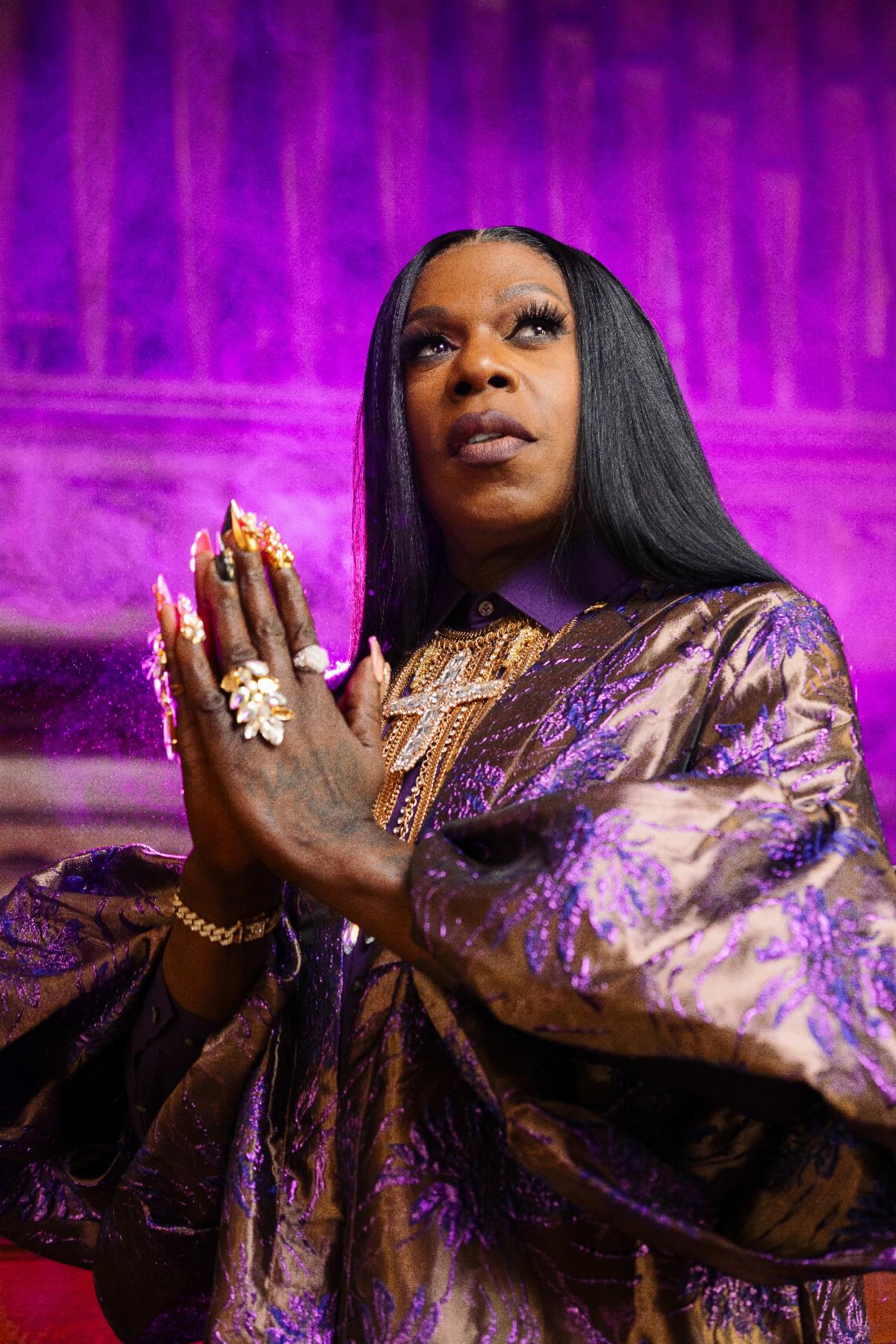 Big Freedia holds her hands together in prayer in a church illuminated by a purple glow.