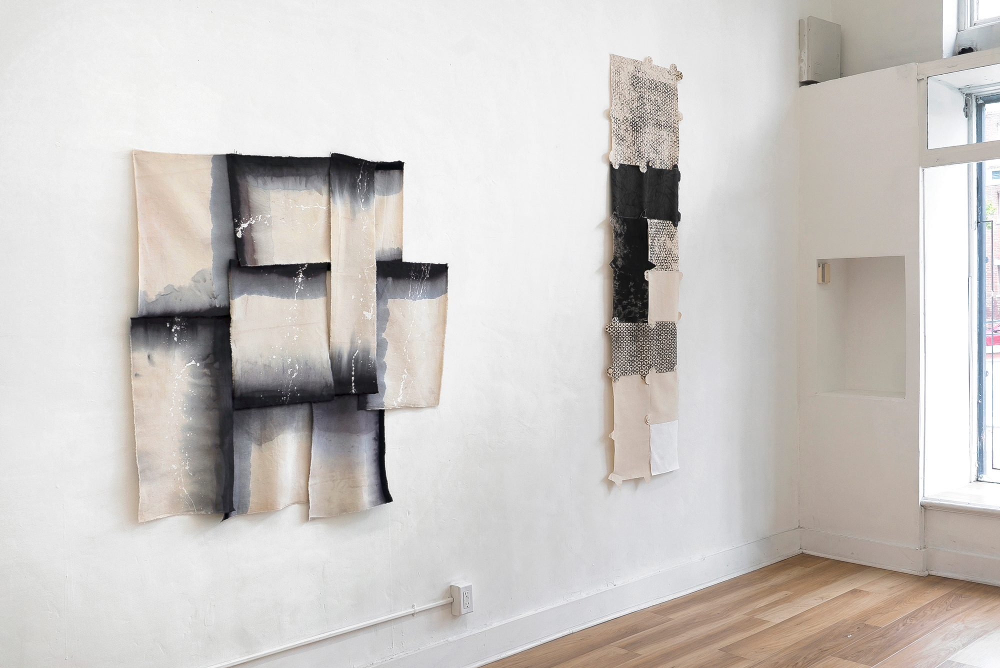 view of corner of gallery with two multi-sectioned textile works in muted grayscale, peek of front window