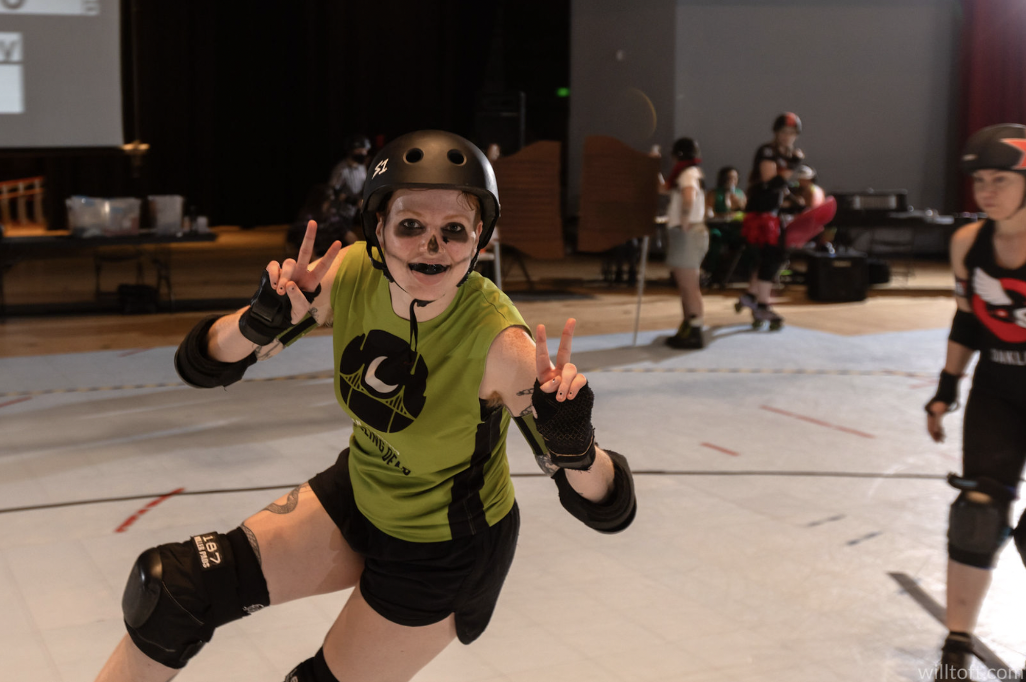 A roller derby skater wearing face paint, knee and elbow pads and a green and black uniform gestures to the camera while skating.